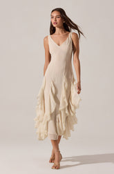 The dress is sleeveless with wide shoulder straps and a V-shaped neckline. It is made of a light, flowing fabric in an off-white or cream color. The dress features vertical seam lines on the bodice and has a layered, ruffled skirt with textured, scalloped edges, creating a tiered effect that extends to mid-calf length. The overall style is elegant and feminine.